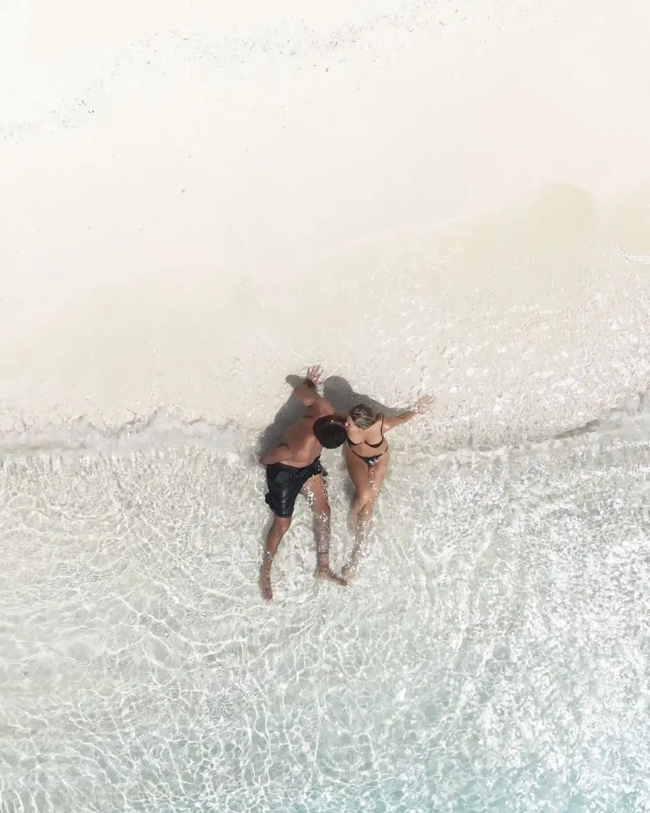 Couple lying in ankle-deep water on a white sandbank, seen from above