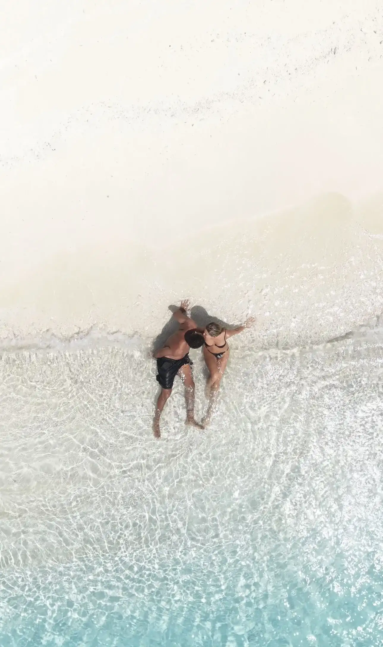 Couple lying in ankle-deep water on a white sandbank, seen from above