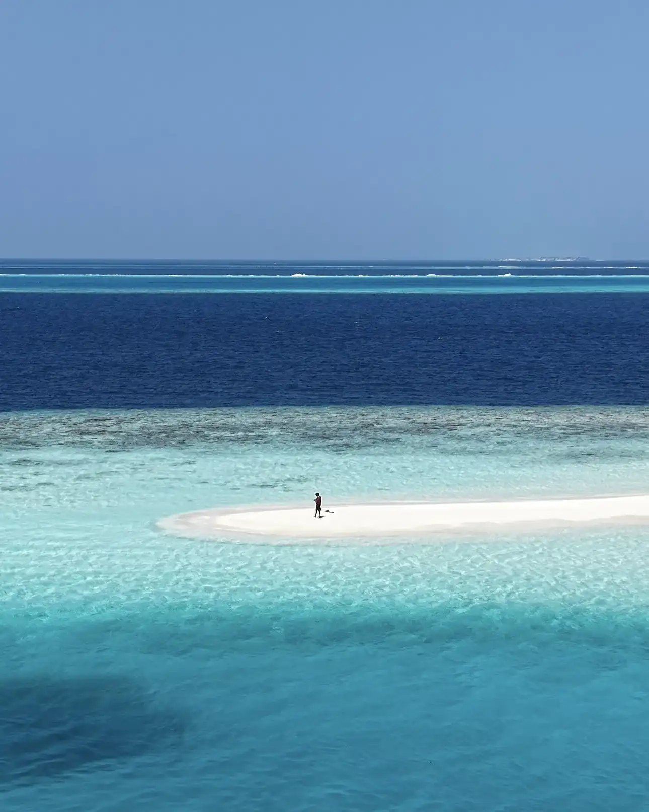 Person standing on a white sandbank between deep blue sea and turquoise shallows