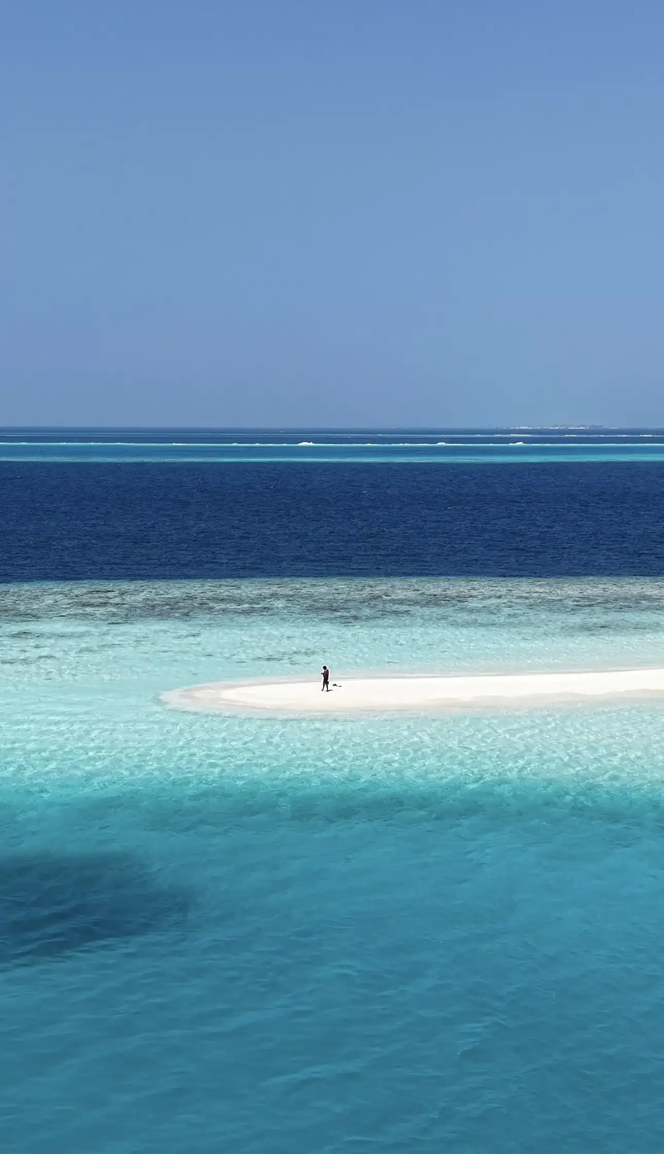 Person standing on a white sandbank between deep blue sea and turquoise shallows